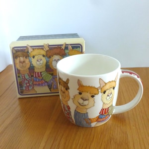 May include: A white ceramic mug featuring a colourful illustration of four alpacas wearing sweaters. The mug has a handle and is in focus. The background is a wooden surface.
