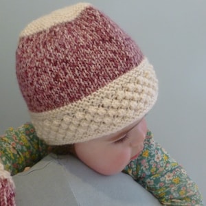 May include: A knitted baby hat with a pink and white color scheme. The hat has a textured pattern on the bottom half.