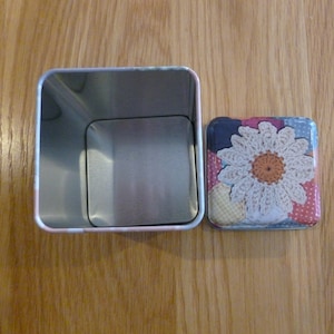 May include: A silver metal tin with a square lid. The lid has a colorful patchwork design with a crocheted white flower in the center.