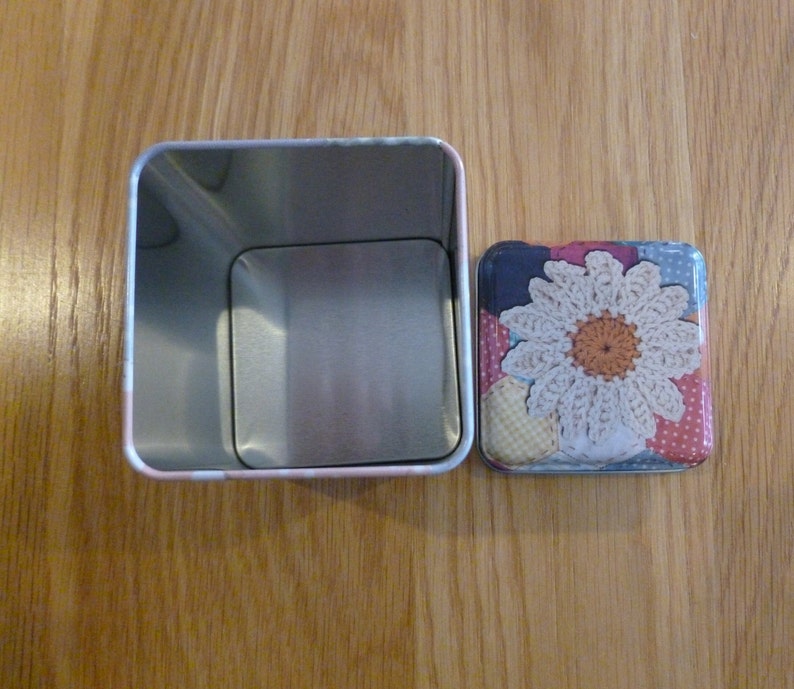 May include: A silver metal tin with a square lid. The lid has a colorful patchwork design with a crocheted white flower in the center.