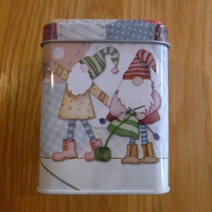 May include: A white metal tin with a colorful patchwork design featuring two gnomes. One gnome is wearing a red and white hat and holding a ball of yarn. The other gnome is wearing a blue and white hat and holding a knitting needle.
