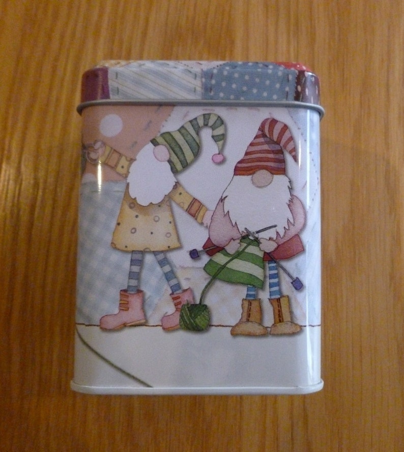 May include: A white metal tin with a colorful patchwork design featuring two gnomes. One gnome is wearing a red and white hat and holding a ball of yarn. The other gnome is wearing a blue and white hat and holding a knitting needle.