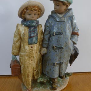 May include: A ceramic figurine depicting two children in winter attire. One child wears a tan coat, hat, and carries a suitcase. The other child is in a blue coat and cap, holding an umbrella. The sculpture has a detailed, hand-painted finish.