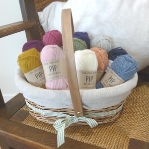 Peut inclure: Un panier en osier rempli de pelotes de laine colorées. La laine est disponible dans des tons de jaune, rose, violet, bleu et crème. Chaque pelote porte une étiquette «PIP» et «100% British Wool». Le panier a une poignée en bois et un ruban rayé vert et blanc.