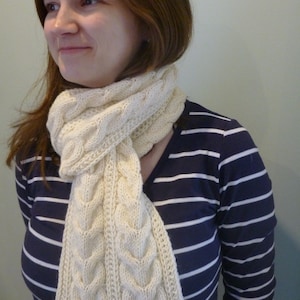 May include: A white knitted cable knit scarf with a loose, flowing design. The scarf is worn by a person with brown hair and a striped shirt.