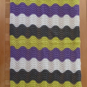 Pode incluir: Um cobertor retangular de malha com um padrão ondulado em tons de verde, roxo, branco e cinza. O cobertor é feito de fio macio e tem uma aparência texturizada. O cobertor tem aproximadamente 100 cm de comprimento e 75 cm de largura.