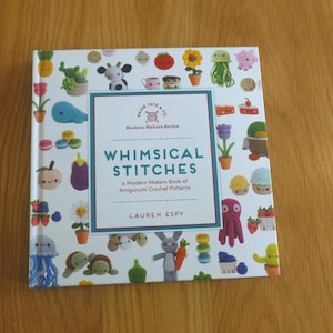 May include: A square hardcover book titled Whimsical Stitches: A Modern Makers Book of Amigurumi Crochet Patterns by Lauren Espy. The white cover features a colorful pattern of various crocheted items including plants, animals, and food.