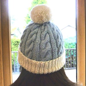 May include: A blue knitted hat with a white pom pom on top. The hat has a white brim and a cable knit pattern.
