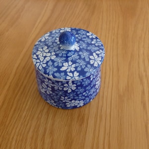 Puede incluir: Un tarro de cerámica azul y blanco con un patrón floral y una tapa con pomo. El tarro está sobre una superficie de madera.