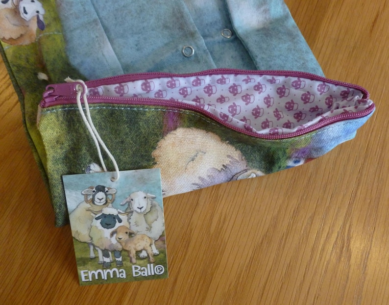 Puede incluir: Un estuche con estampado de ovejas y cremallera morada. El forro interior presenta un patr&oacute;n repetido de peque&ntilde;as ovejas. Una etiqueta con el texto "Emma Ball&reg;" y una ilustraci&oacute;n de ovejas est&aacute; adjunta.