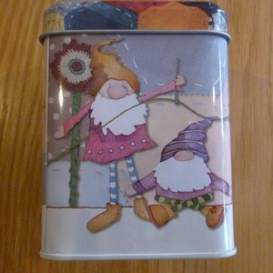 May include: A white metal tin with a colorful patchwork design featuring two gnomes. One gnome is holding a needle and thread, while the other gnome is wearing a purple hat and green pants.