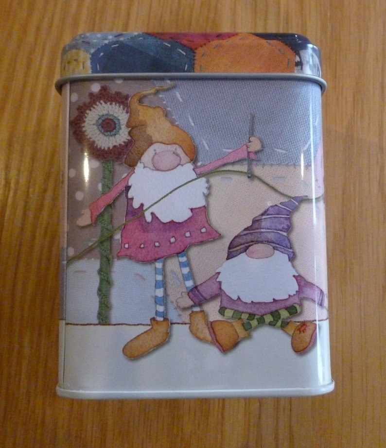May include: A white metal tin with a colorful patchwork design featuring two gnomes. One gnome is holding a needle and thread, while the other gnome is wearing a purple hat and green pants.