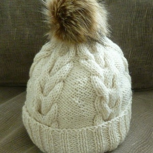 May include: A white knitted beanie with a brown faux fur pom pom. The beanie has a cable knit pattern.