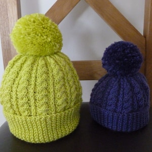 May include: Two knitted bobble hats. One is lime green, the other is dark purple. Both have a cable knit pattern and a ribbed brim. The hats are displayed on a dark surface.