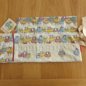 Puede incluir: Dos estuches rectangulares de tela crema con un estampado de ovejas con coloridos su&eacute;teres. Uno de los estuches est&aacute; abierto, mostrando un dise&ntilde;o tipo regla. Tambi&eacute;n hay una etiqueta con una ilustraci&oacute;n de oveja y una cinta crema.
