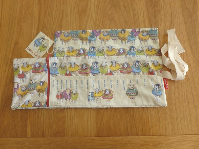 Puede incluir: Dos estuches rectangulares de tela crema con un estampado de ovejas con coloridos su&eacute;teres. Uno de los estuches est&aacute; abierto, mostrando un dise&ntilde;o tipo regla. Tambi&eacute;n hay una etiqueta con una ilustraci&oacute;n de oveja y una cinta crema.