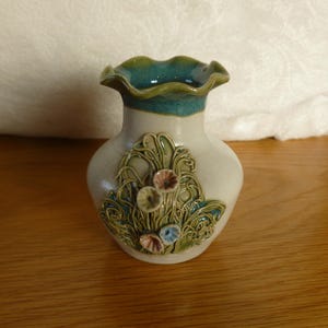 May include: A small, white ceramic vase with a scalloped rim and a green glaze. The vase has a raised design of flowers and leaves.