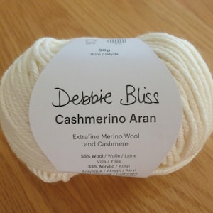 以下が含まれることがあります： Debbie Bliss Cashmerino Aranと書かれた紙ラベルが付いたオフホワイトの毛糸玉。ラベルにはExtra-fine Merino Wool and Cashmere、50g、90m、ウール55%、アクリル33%、カシミヤ12%と記載されています。毛糸は太く、柔らかくねじれた質感です。