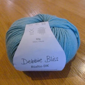 Op de afbeelding: Een bol lichtblauw garen met een label "Debbie Bliss Rialto DK". Het label geeft 50g en 105m aan. Het garen is een zacht, getextureerd materiaal, ideaal voor brei- of haakprojecten. Het label geeft ook 4mm US 6 naalden aan.