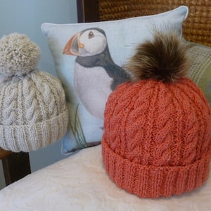 May include: Two knitted hats with pom poms. One hat is beige with a white pom pom and the other is orange with a brown fur pom pom.
