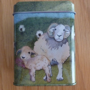May include: A green tin with a white rim features a watercolor illustration of a brown and white sheep and a smaller lamb standing in a grassy field. The sheep has large curled horns and the lamb has a white face.
