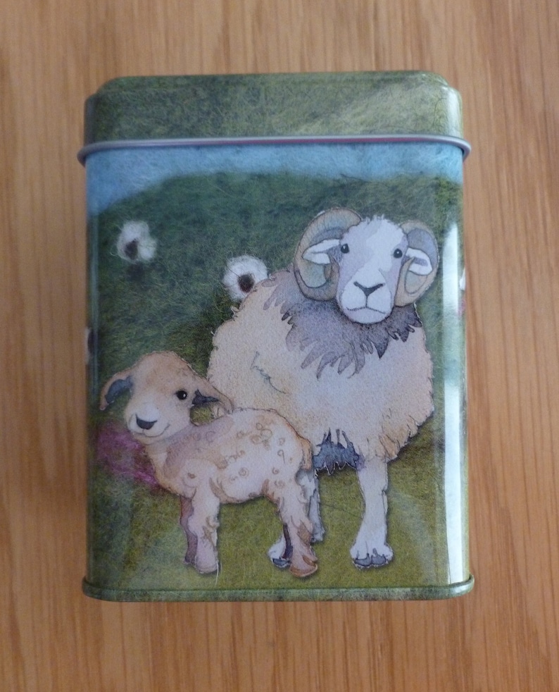 May include: A green tin with a white rim features a watercolor illustration of a brown and white sheep and a smaller lamb standing in a grassy field. The sheep has large curled horns and the lamb has a white face.