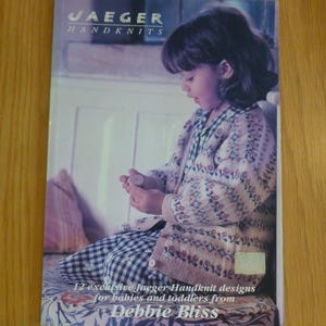 May include: A book cover featuring the text "JAEGER HANDKNITS" and a photograph of a child in a floral cardigan and checkered trousers. The book's title is "12 exclusive Jaeger Handknit designs for babies and toddlers from Debbie Bliss."