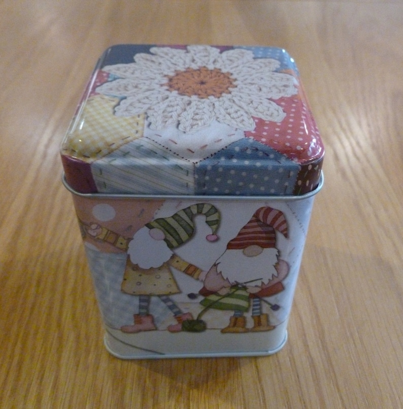 May include: A square tin box with a patchwork design featuring a white crocheted flower. The box has a colorful design with gnomes knitting on the front.