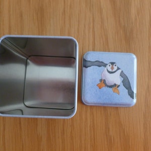 May include: A square metal tin with a removable lid. The lid features a colorful illustration of a puffin with orange feet and a blue background.