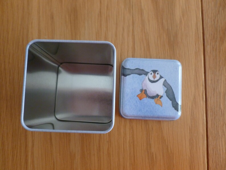 May include: A square metal tin with a removable lid. The lid features a colorful illustration of a puffin with orange feet and a blue background.