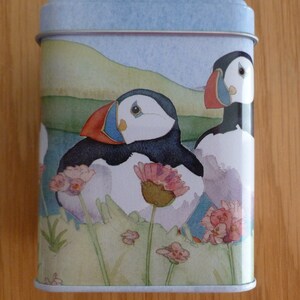 May include: A blue and white tin with a watercolor illustration of two puffins on a grassy cliff with pink flowers. The puffins are facing the viewer with their beaks open.