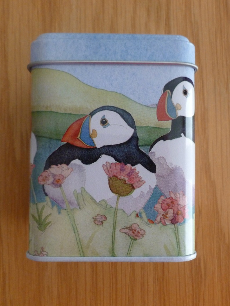 May include: A blue and white tin with a watercolor illustration of two puffins on a grassy cliff with pink flowers. The puffins are facing the viewer with their beaks open.