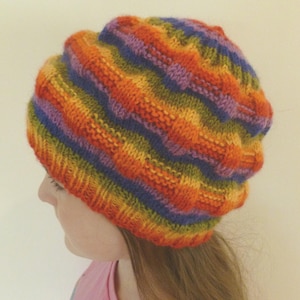 May include: A knit beanie featuring a vibrant, multi-colored horizontal striped pattern in shades of orange, yellow, green, purple, and blue. The hat has a textured, ribbed stitch design that creates a raised, wavy appearance across the fabric.