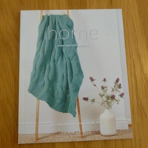 May include: Book cover with the title "home" in white text. The cover features a teal knitted blanket draped over a wooden stand. The book is titled "home: Seven hand knit patterns" and is published by John Lewis.