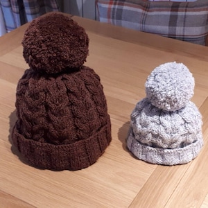 Könnte beinhalten: Zwei Strickmützen mit Bommeln. Die größere Mütze ist braun mit einem Zopfmuster. Die kleinere Mütze ist grau mit einem Zopfmuster.