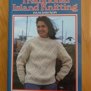 May include: A book titled "Traditional Island Knitting" by Pam Dawson. The cover features a woman wearing a white knitted sweater with a diamond pattern, standing in front of a harbor scene.