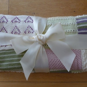 May include: A knitted baby blanket with a patchwork design in shades of pink, green, and white. The blanket is tied with a white satin ribbon.
