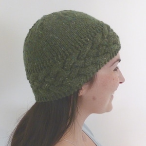 May include: A forest green knit beanie featuring a textured cable knit pattern around the brim and a smooth stockinette stitch on the crown. The yarn has subtle flecks of lighter green and brown throughout the dark green base color.