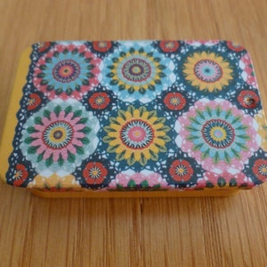 May include: A small, rectangular tin box with a colorful floral pattern. The tin has a yellow background with a repeating pattern of blue, pink, and yellow flowers.