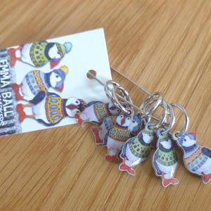 May include: A set of six stitch markers on a silver-toned safety pin, attached to a white card labelled Emma Ball Six Stitch Markers. Each marker features a small, colourful illustration of a puffin wearing a patterned jumper and a knit hat.