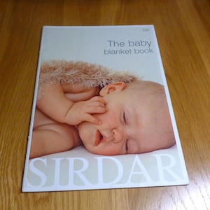 Puede incluir: Un libro titulado "The baby blanket book" con una foto de un bebé durmiendo en la portada. El bebé tiene la piel clara y descansa sobre una superficie blanca, con una manta suave y marrón. La marca "SIRDAR" está impresa en la parte inferior.