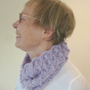Könnte beinhalten: Ein helllila gestrickter Rundschal mit einem strukturierten Muster. Der Schal wird um den Hals getragen und hat einen gerüschten Rand. Die Person auf dem Bild trägt ein weißes Hemd und eine Brille.