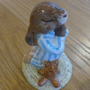 Può includere: Una statuetta in ceramica di un coniglio marrone che indossa un pigiama blu e bianco, tenendo un orsacchiotto di peluche marrone. Il coniglio è in piedi su una base color crema.