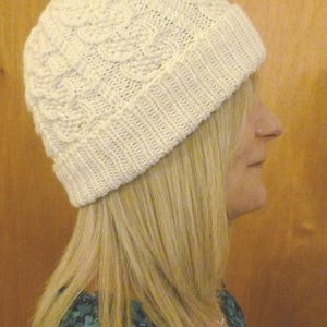 May include: A white knitted hat with a cable knit pattern. The hat is worn by a person with long blonde hair.