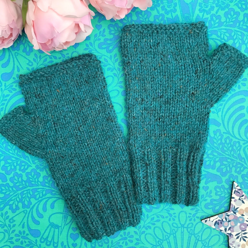May include: A pair of teal blue fingerless gloves made of a soft, textured knit fabric. The gloves are shown on a patterned background.