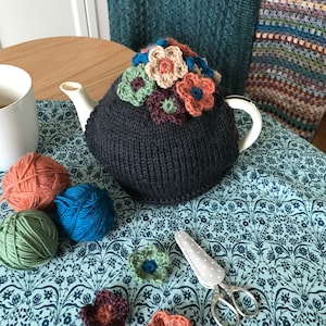 May include: A navy blue knitted tea cosy decorated with crocheted flowers in various colours. The cosy is on a floral tablecloth, with balls of yarn in orange, blue, and green nearby. A pair of scissors with a grey polka-dot handle is also visible.