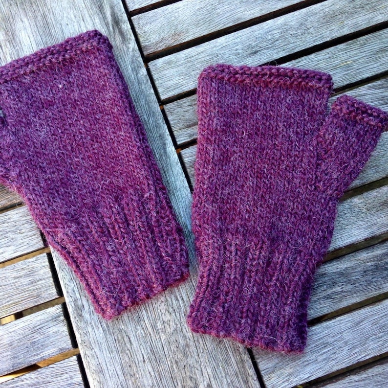 May include: A pair of purple fingerless gloves made of knitted wool. The gloves are laying on a wooden surface.