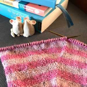 May include: Close-up of a pink and white striped knitted fabric with a knitting needle in the fabric. The fabric is on a gray surface with two ceramic mice figurines in the background.