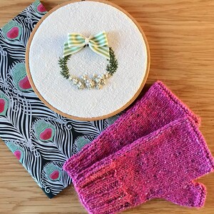 May include: A pair of pink knitted fingerless gloves with a white embroidery hoop with a green and white striped bow and a floral wreath.