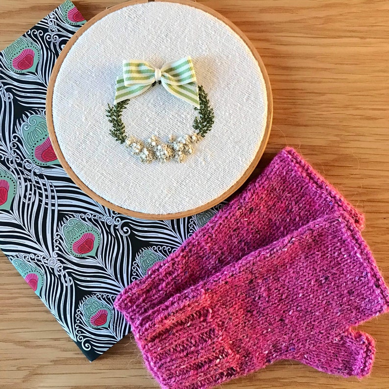 May include: A pair of pink knitted fingerless gloves with a white embroidery hoop with a green and white striped bow and a floral wreath.